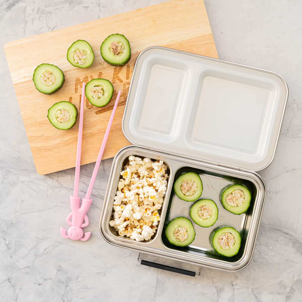 Tuna and Rice mixture stuffed inside hollowed out cucumber pieces on a chopping board, a childs lunchbox with Cucumber Sushi and popcorn inside.