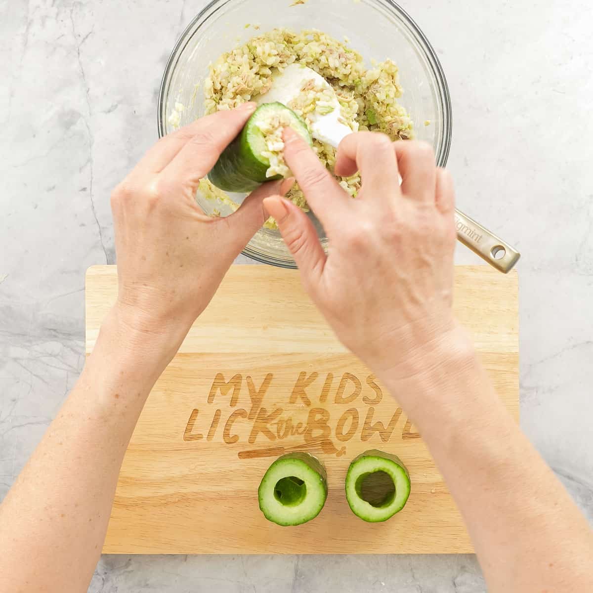 Hand holding a piece of hollowed out Cucumber over the bowl of tuna rice mixture which is being stuffed into the cucumber. Two hollowed cucumbers on chopping board also.