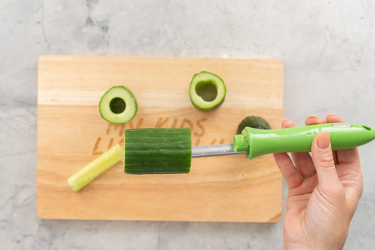 Hand holding an apple corer with a cucumber piece on the end hallowing out the centre. Hollowed cucumber pieces on chopping board.