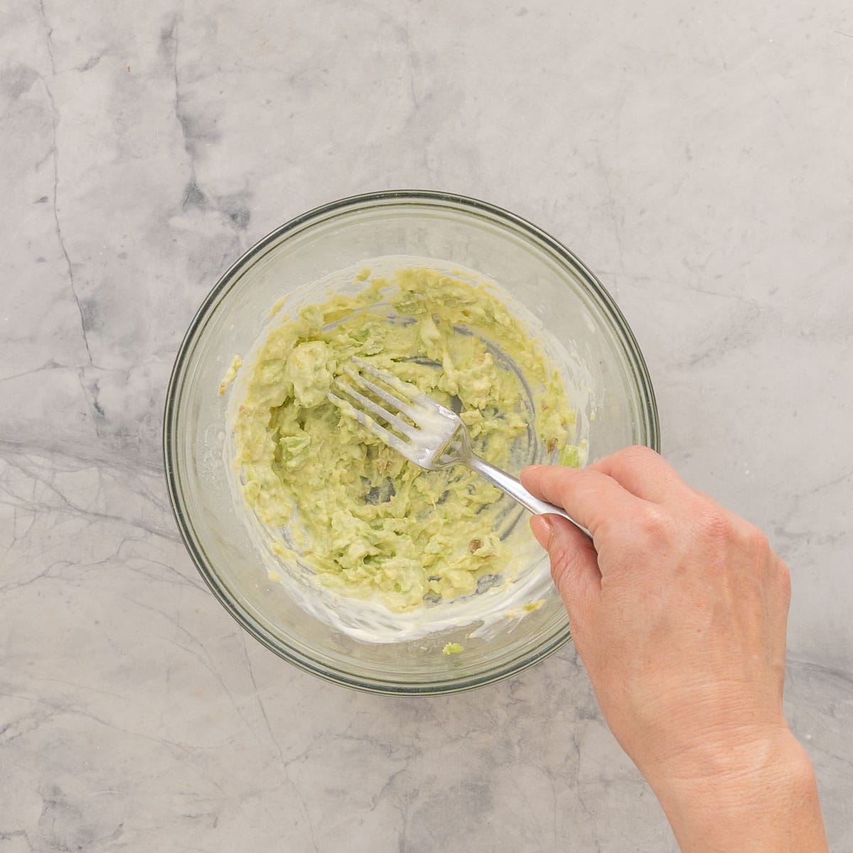 Avocado, mayonnaise, tuna and lemon juice in a glass bowl being mashed together with a fork.