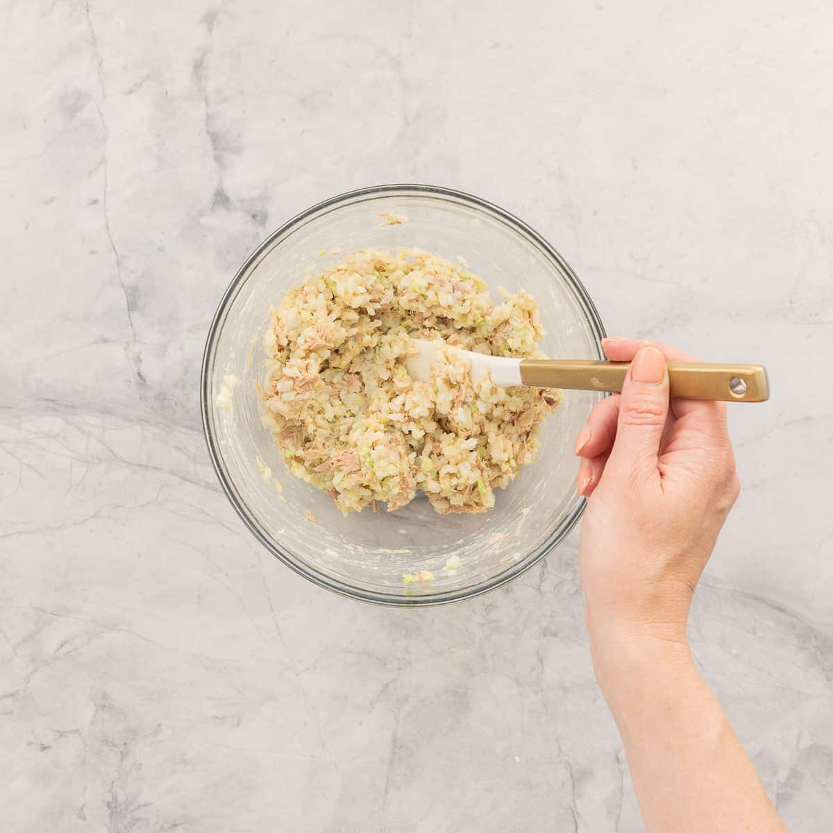 Avocado, mayonnaise, tuna and lemon juice and cooked rice in a glass bowl being mixed with a spatula.