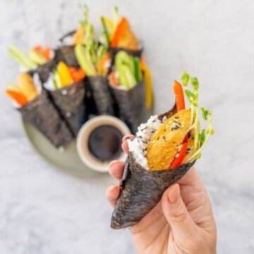 Hand holding a piece of Hand roll sushi with Ingredients coming out the top garnished with sesame seeds.