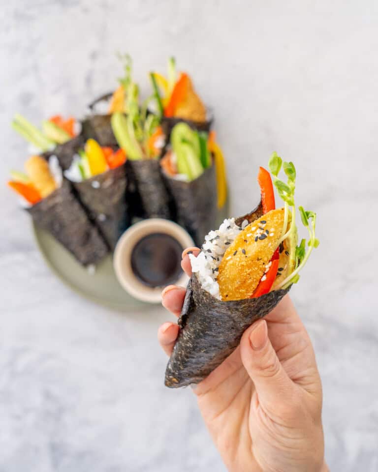 Hand holding a piece of Hand roll sushi with Ingredients coming out the top garnished with sesame seeds.