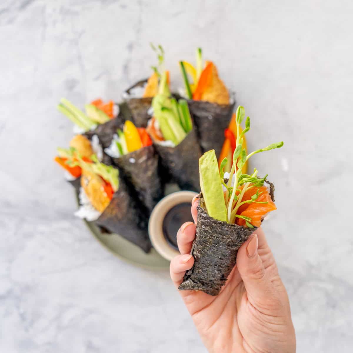 Hand roll sushi piece being held up with rest on a plate with dish of soy sauce blurred in background.