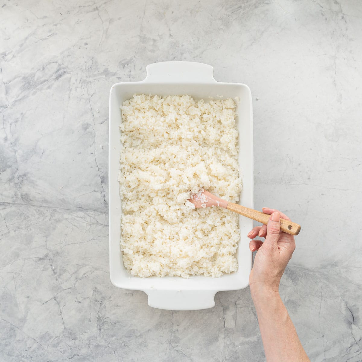 Hand using a spatula to spread out Cooked sushi rice in a shallow dish.