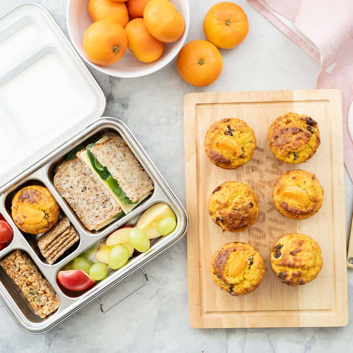 Mandarin muffins cooked in silicon muffin tray on bench top with a lunchbox filled with mandarin muffin, sandwich, cherry tomatoes, muesli bar, apples and grapes.