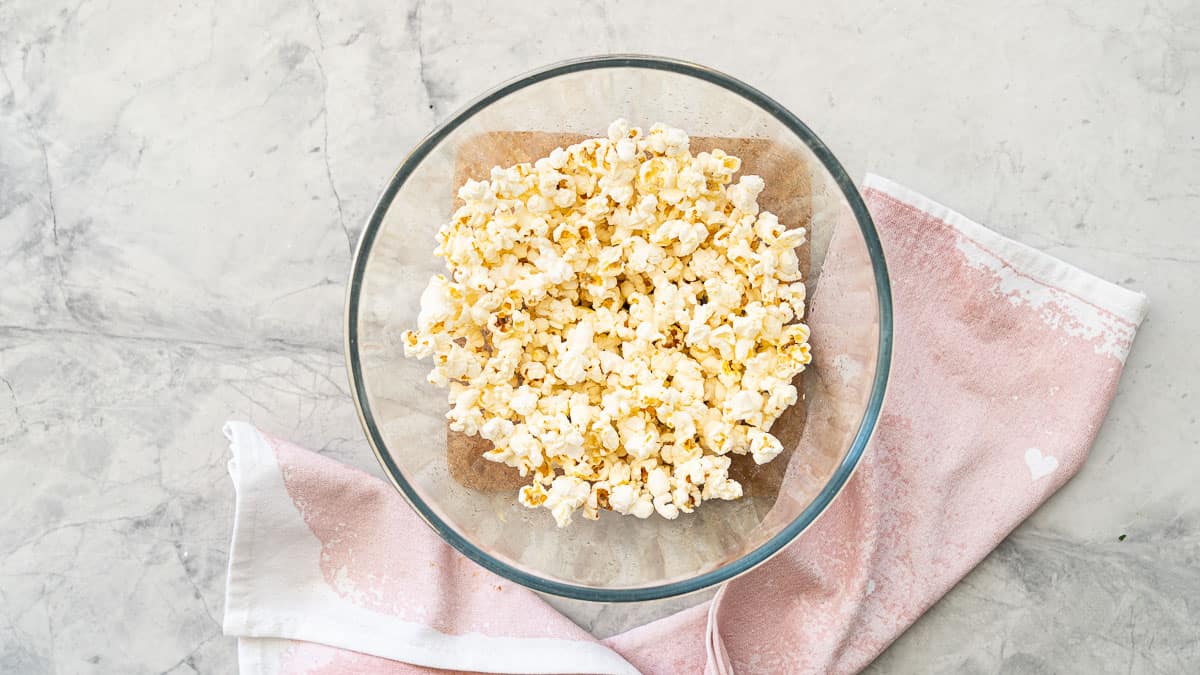 Large glass bowl on bench top with popcorn inside next to a dusky pink hand towel.