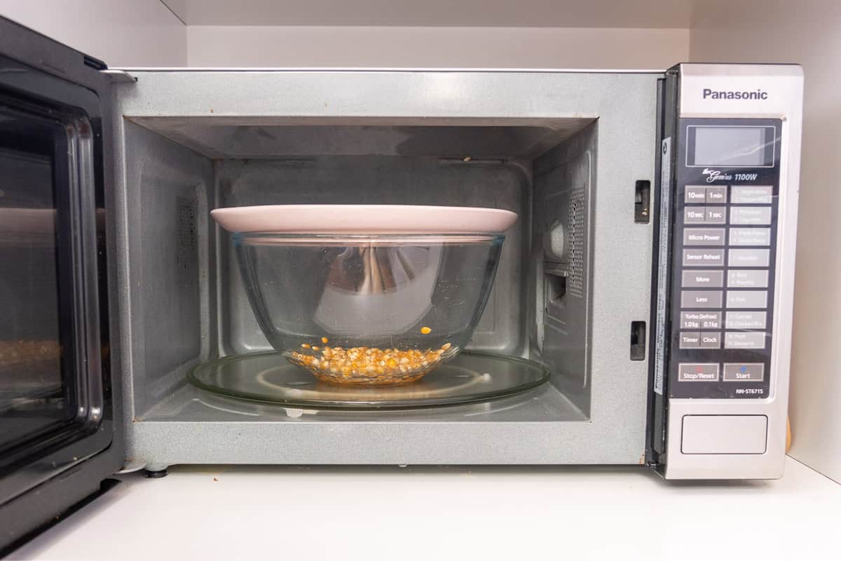 Large glass bowl in microwave with plate covering the top, with popcorn kernels and coconut oil inside.