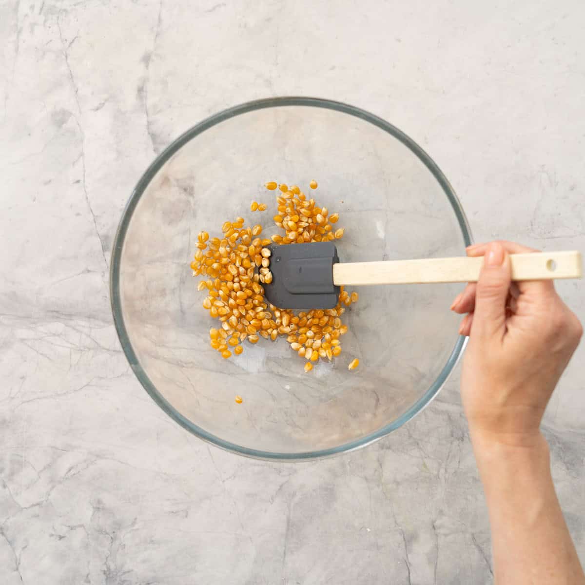 Melted Coconut oil and popcorn kernels in a large glass bowl on benchtop being mixed with spatula.