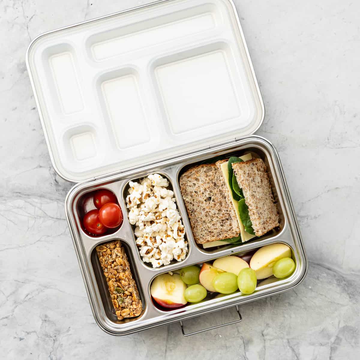 Lunchbox with assortment of food inside, popcorn, sandwich, cherry tomatoes, muesli bar, apples and grapes.