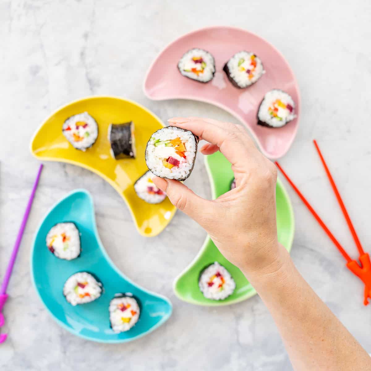 Hand holding a piece of rainbow sushi up to camera above four moon shaped colourful plates on bench top with rainbow sushi showing ingredients inside.