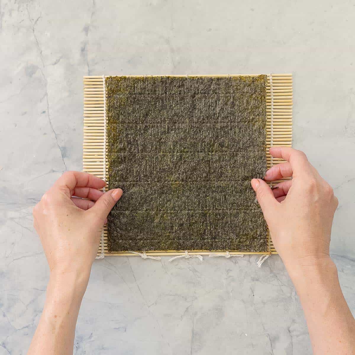 Sushi rolling mat on bench top, hands placing a piece of Sushi Paper on mat.