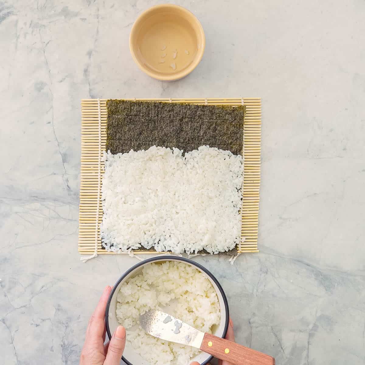 Sushi rolling mat on bench top with sushi paper on top and thin layer of rice covering ¾ of sushi paper. Bowl of cooked rice with flat metal spatula inside and small ramekin of water on the benchtop.