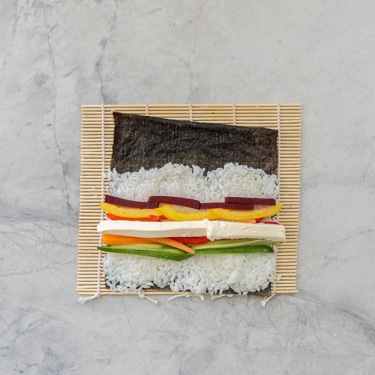 Sushi mat on bench top with Sushi paper and thin layer of sushi rice, Thin sticks of Cucumber, carrot, red and yellow capsicum, beetroot.