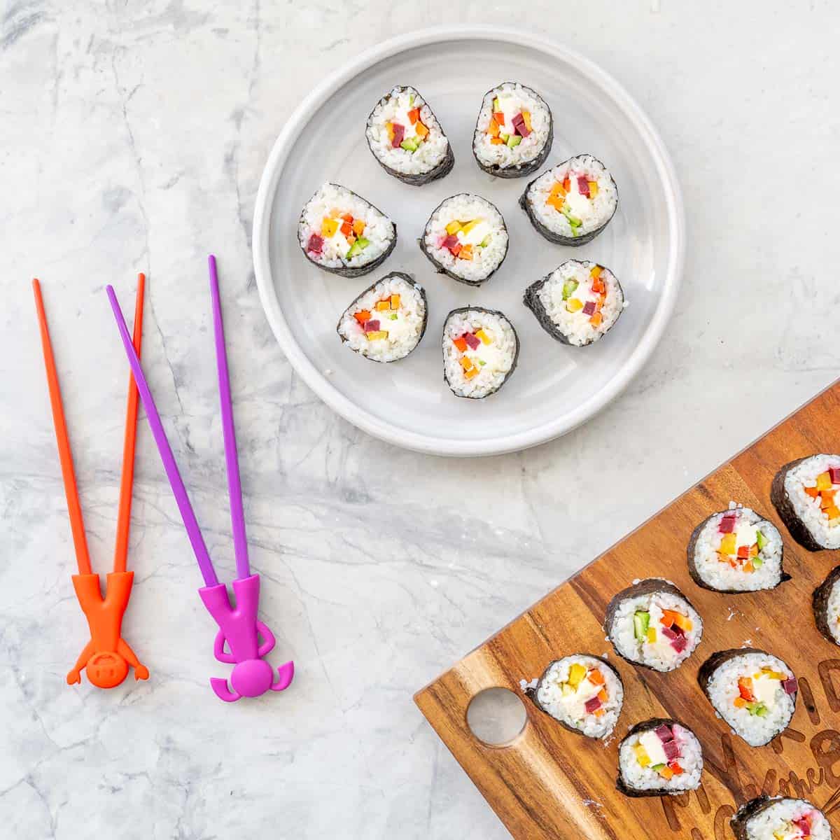 Sushi roll cut into pieces with ingredients showing inside, placed on a plate and wooden chopping board. Children's chopsticks on benchtop.