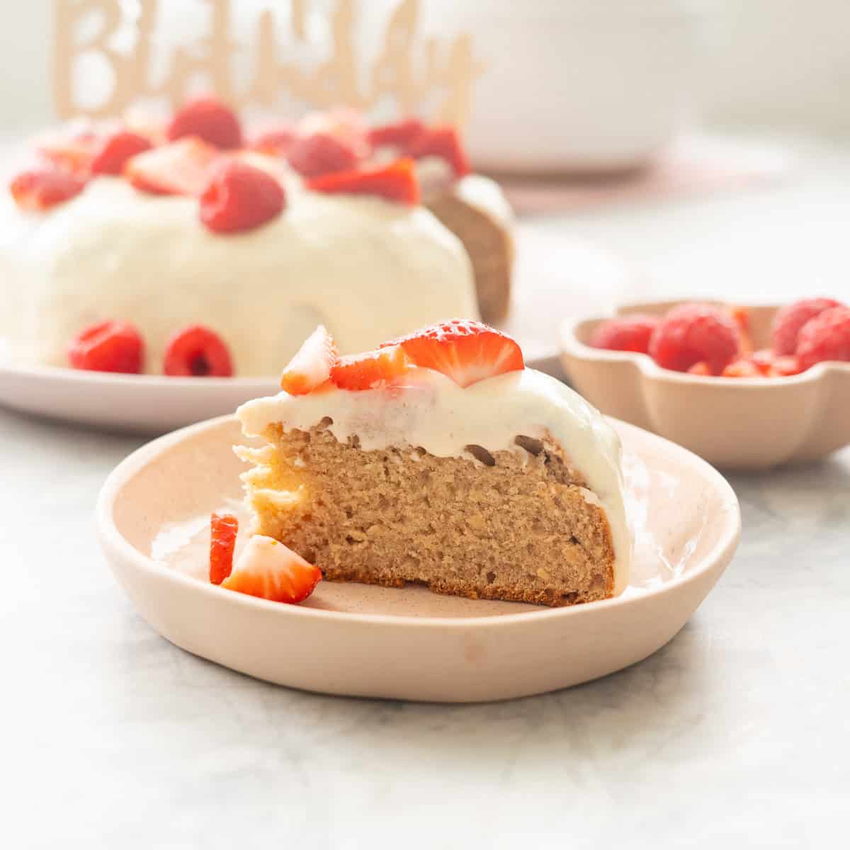 Slice of Smash Cake served on a blush pink plate, topped with frosting and garnished with halved strawberries, raspberries. Cake blurred in background topped with "Happy birthday" sign.