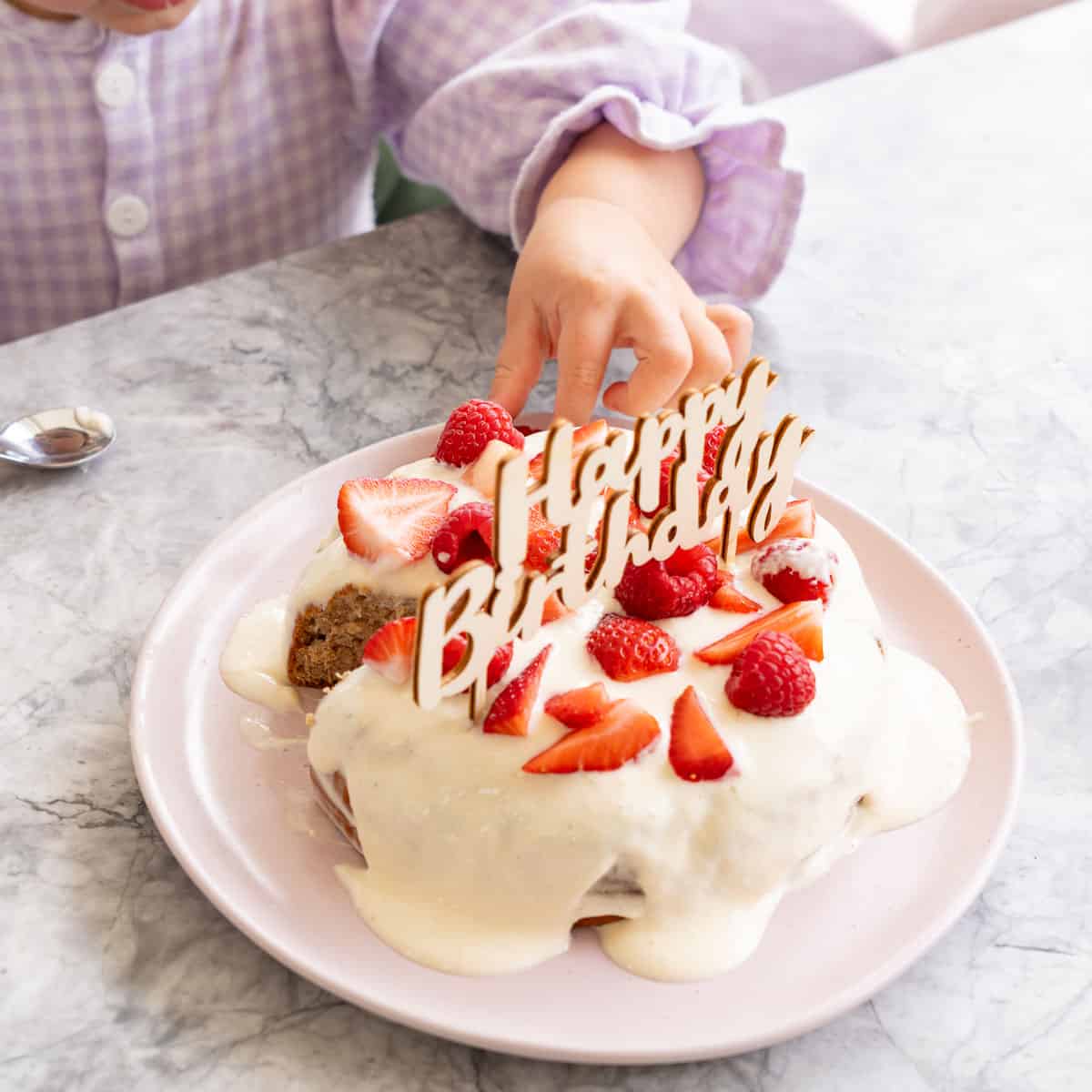 Smash Cake served on a blush pink plate, topped with frosting and garnished with halved strawberries, raspberries and topped with "Happy birthday" sign, babies had grading a strawberry off the top of the cake.