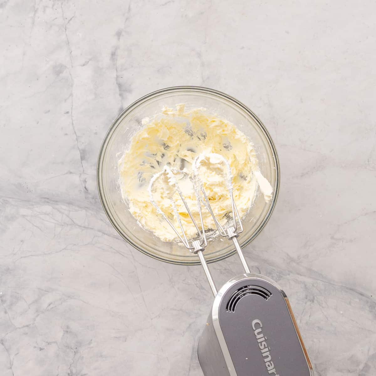 Cream cheese in a glass bowl on bench top being whisked with an electric beater.