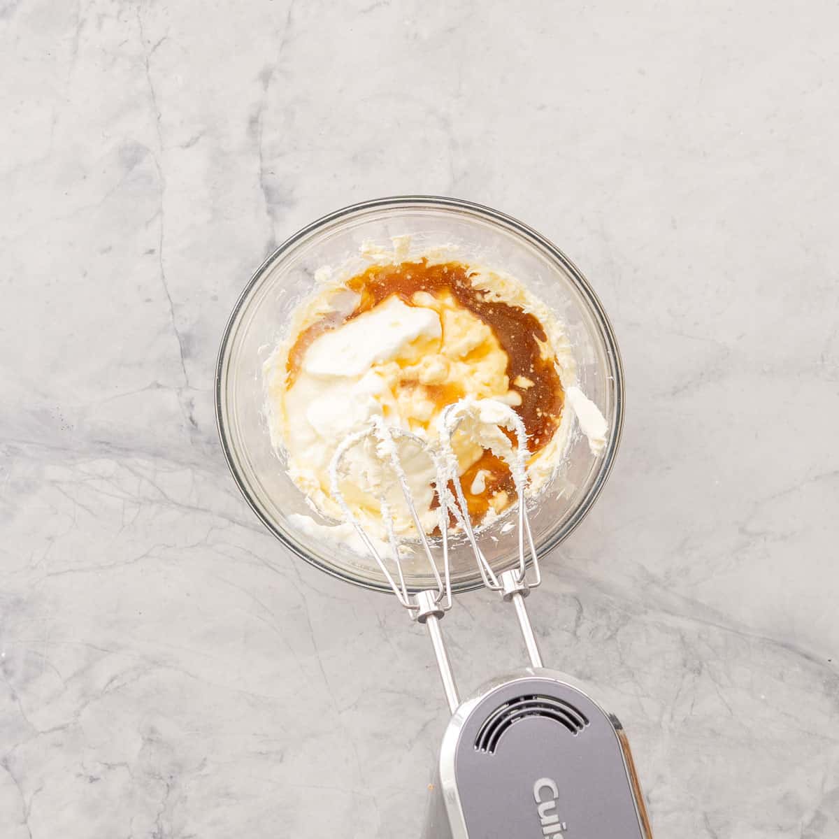 Ingredients for Smash Cake added into whipped cream cheese in a glass bowl on bench top being whisked with an electric beater.