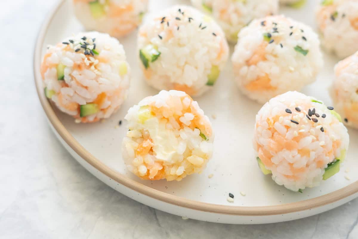 Sushi rice mixture rolled into balls on a plate with sesame seeds garnished on top. One ball bit in half showing cream cheese inside.