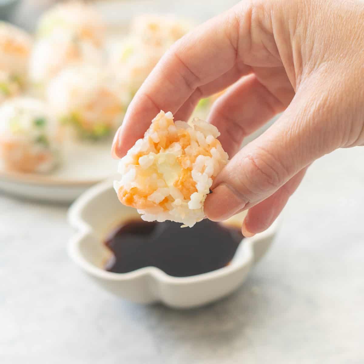 Hand holding half of Sushi ball showing cream cheese inside, being dipped into soy sauce with rest of balls on a plate with sesame seeds garnished on top blurred in background.