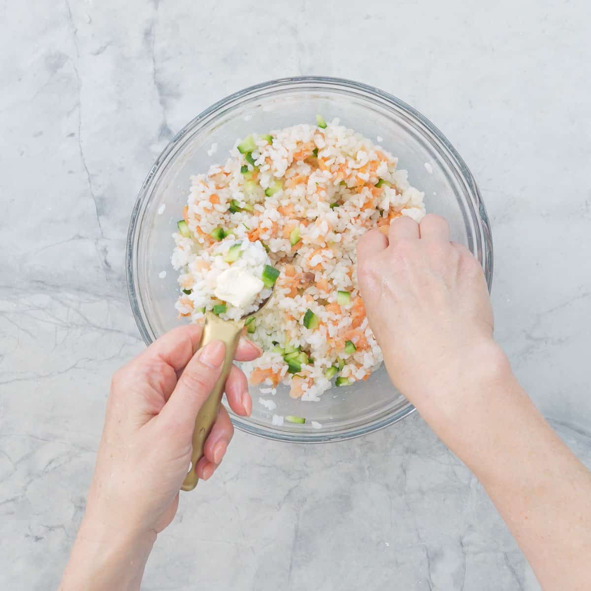 Sushi ball mixture in big glass bowl, hand holding a spoon with scoop of mixture and cream cheese on top.