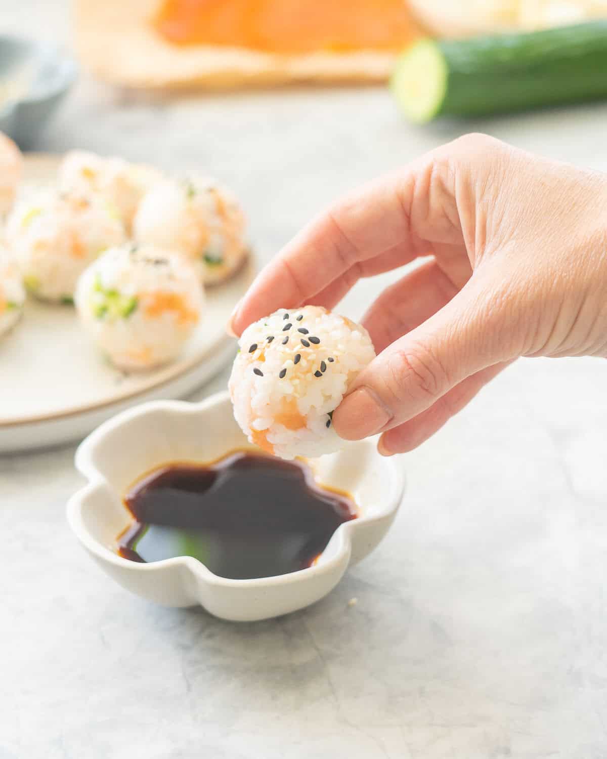 Hand holding Sushi ball being dipped into soy sauce, rest of balls on a plate with sesame seeds garnished on top blurred in background.