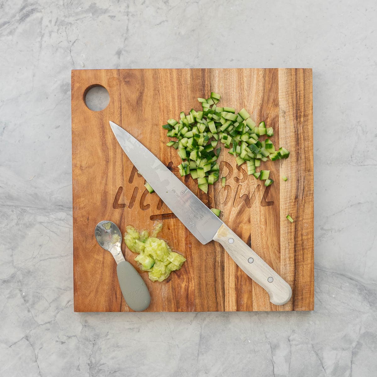 Knife laid across a wooden chopping board next to diced cucumber pieces and core of cucumber mashed next to baby spoon.