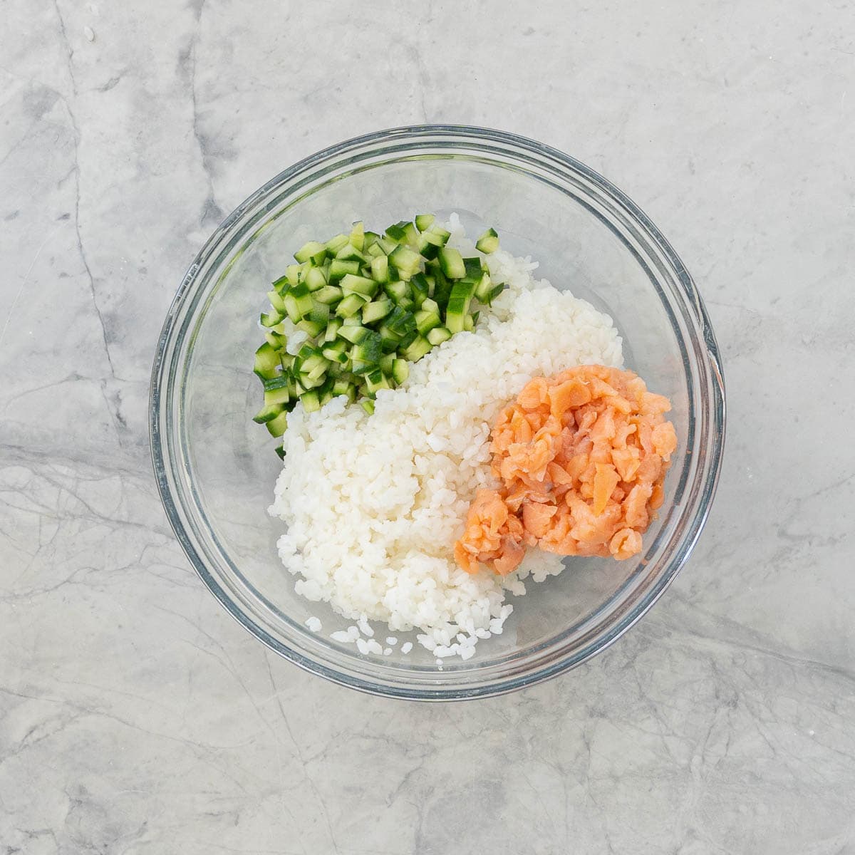 Diced cucumber, cooked sushi rice and diced salmon pieces in a big glass bowl.