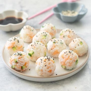 Sushi rice mixture rolled into balls on a plate with sesame seeds garnished on top with soy sauce and chopsticks blurred in background.