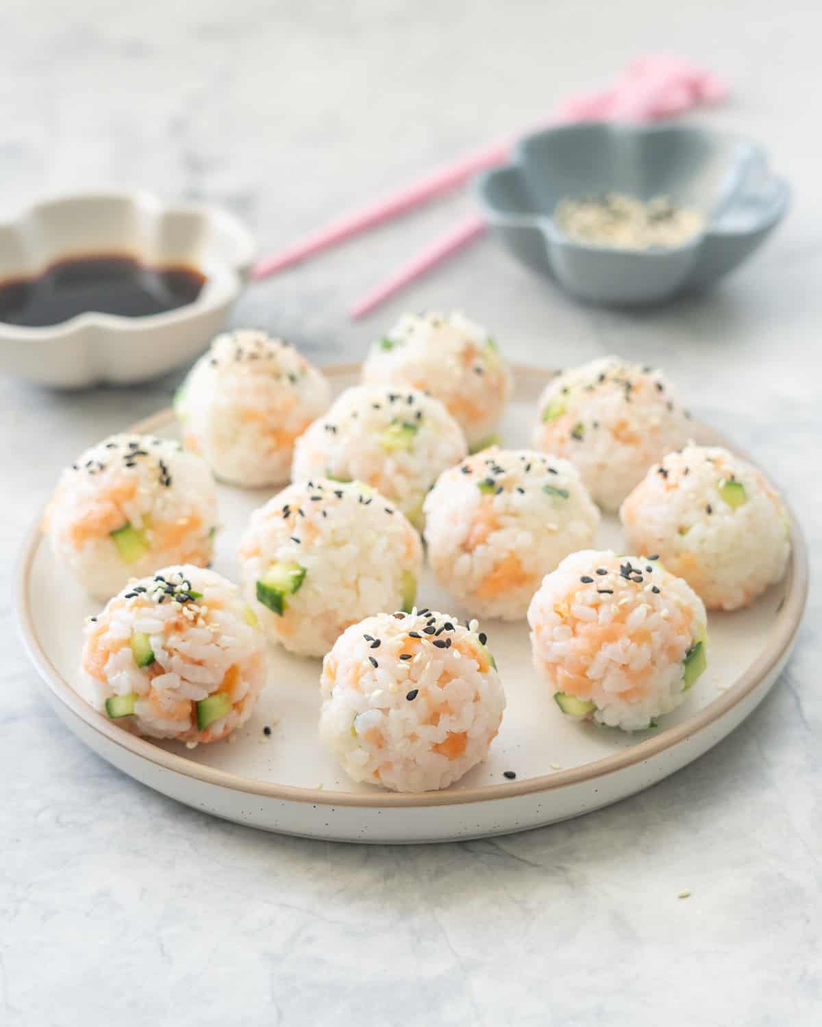 Sushi rice mixture rolled into balls on a plate with sesame seeds garnished on top with soy sauce and chopsticks blurred in background.