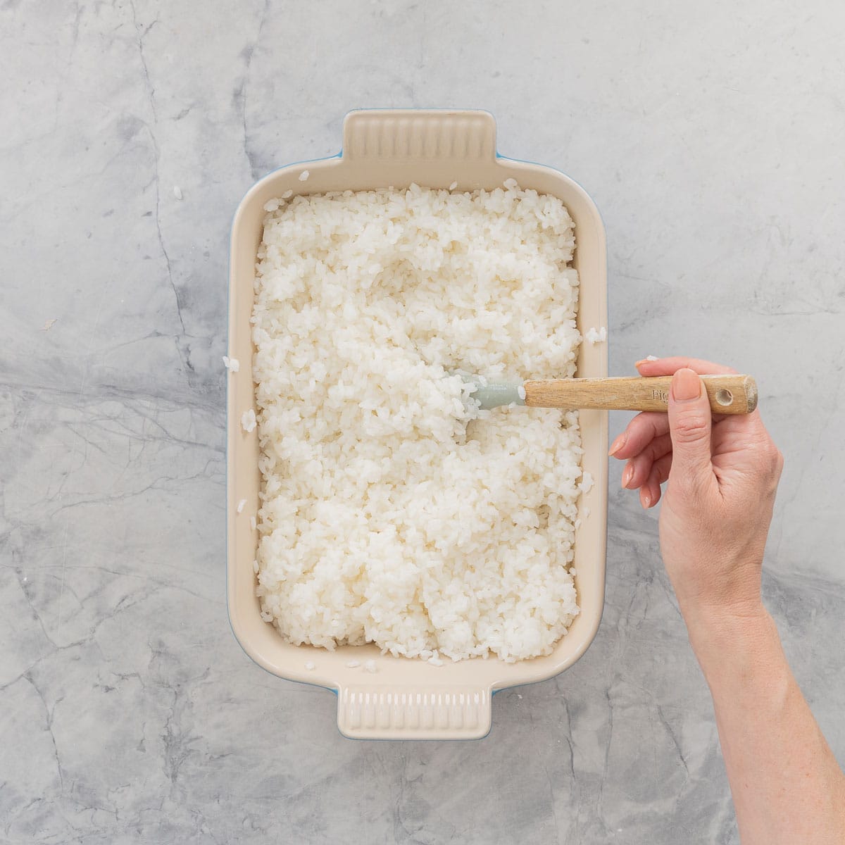 Hand using a spatula to spread out Cooked sushi rice in a shallow dish.