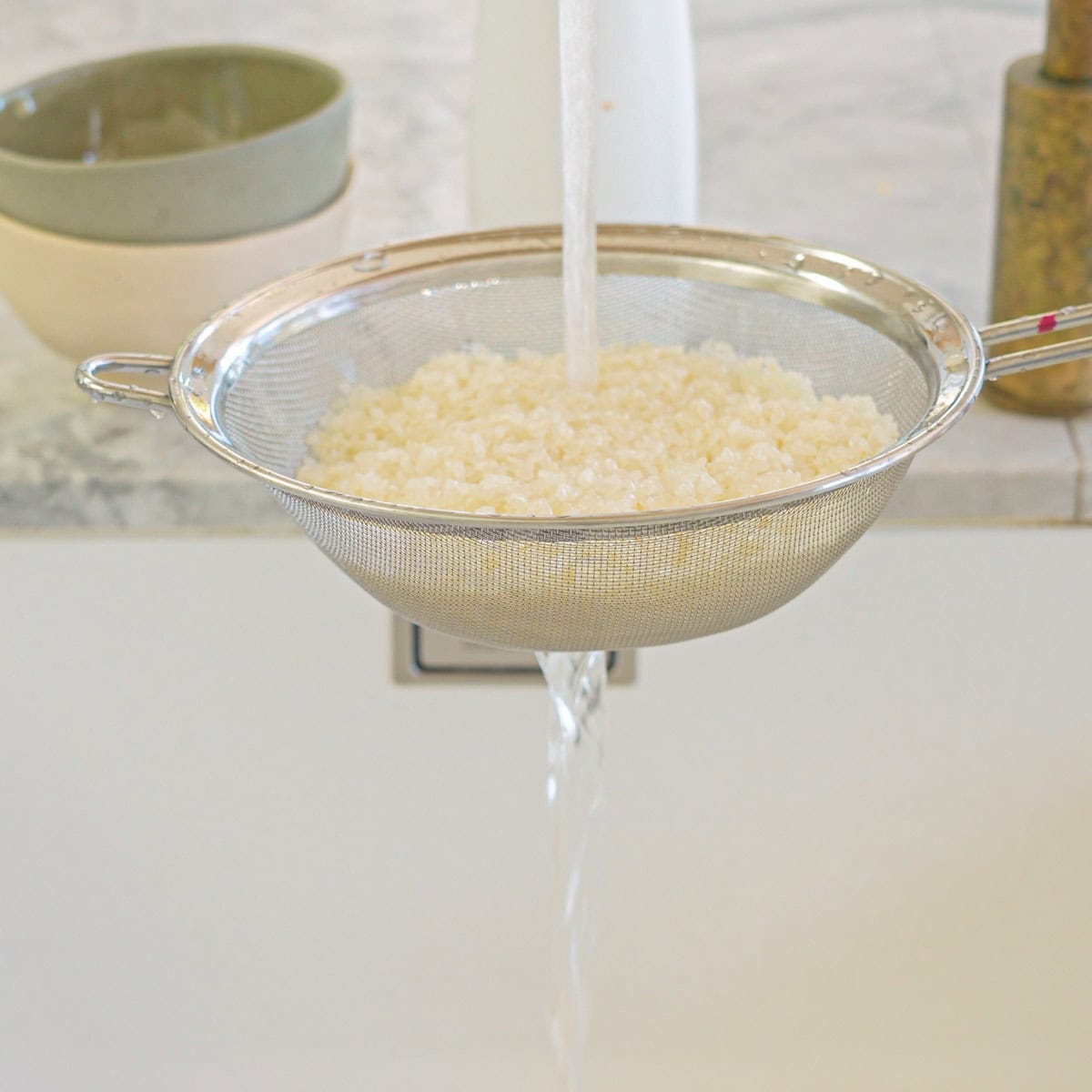 Sushi rice in fine mesh strainer being rinsed under cold water.