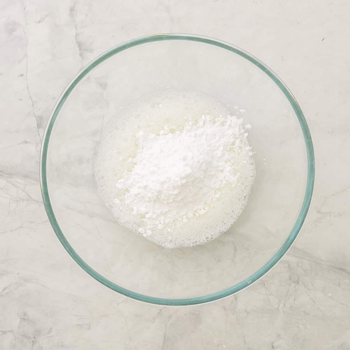 Whisked egg whites and icing sugar in a glass bowl on benchtop.