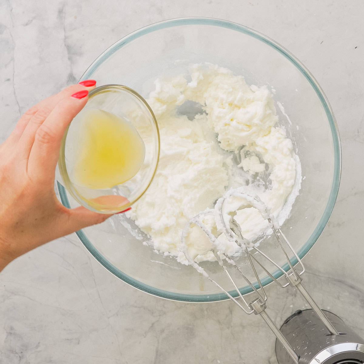 Hand holding a small glass bowl pouring lemon juice into whisked icing sugar and egg whites in big glass bowl.