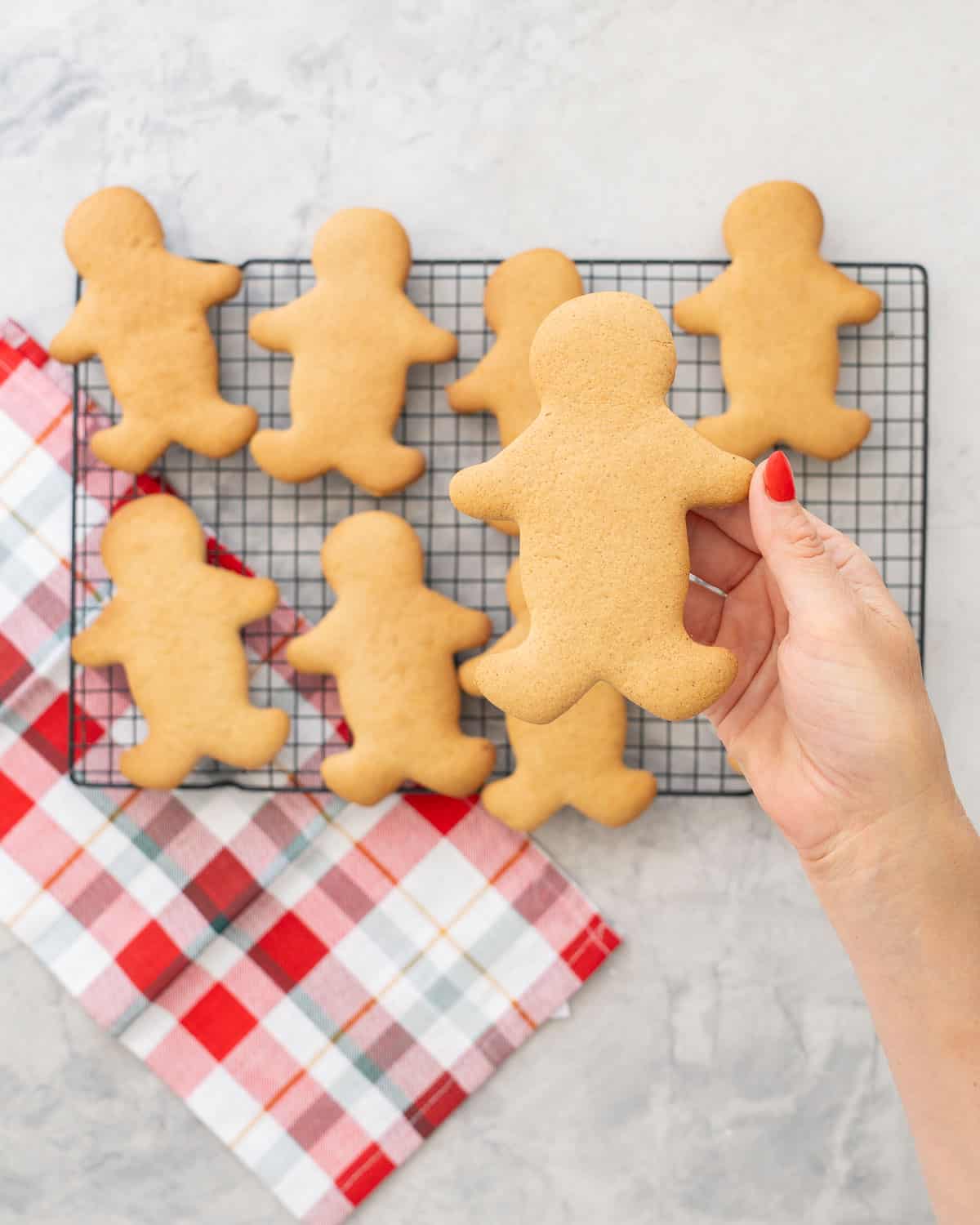 Hand holding up a cooked gluten free gingerbread man to camera, cooling tray on bench in background with batch of gingerbread.