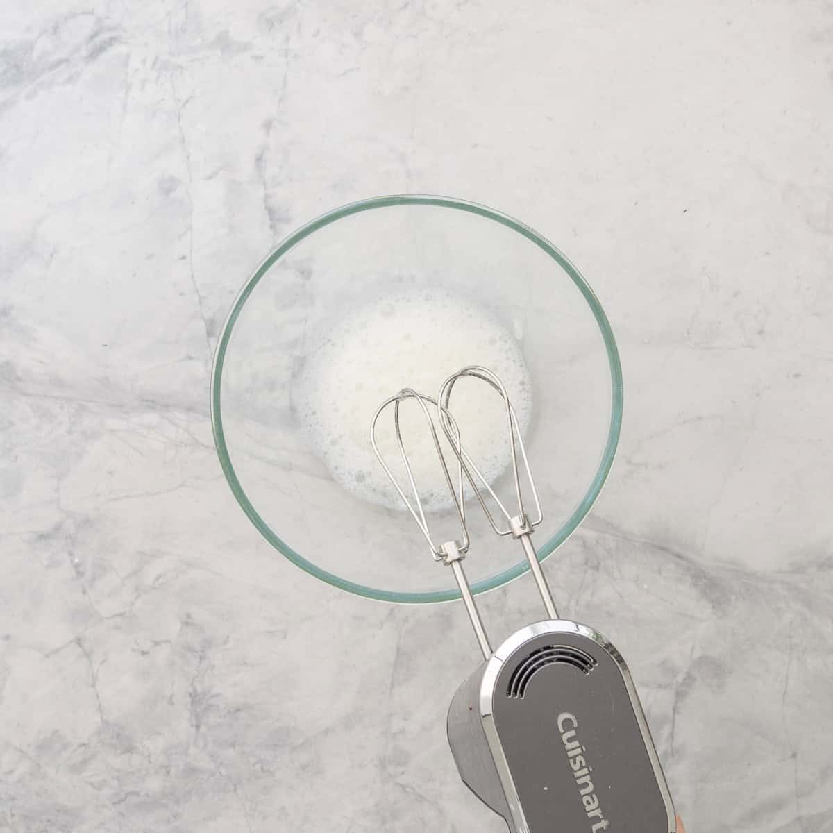 Egg whites whisked in glass bowl on bench top with electric beater.