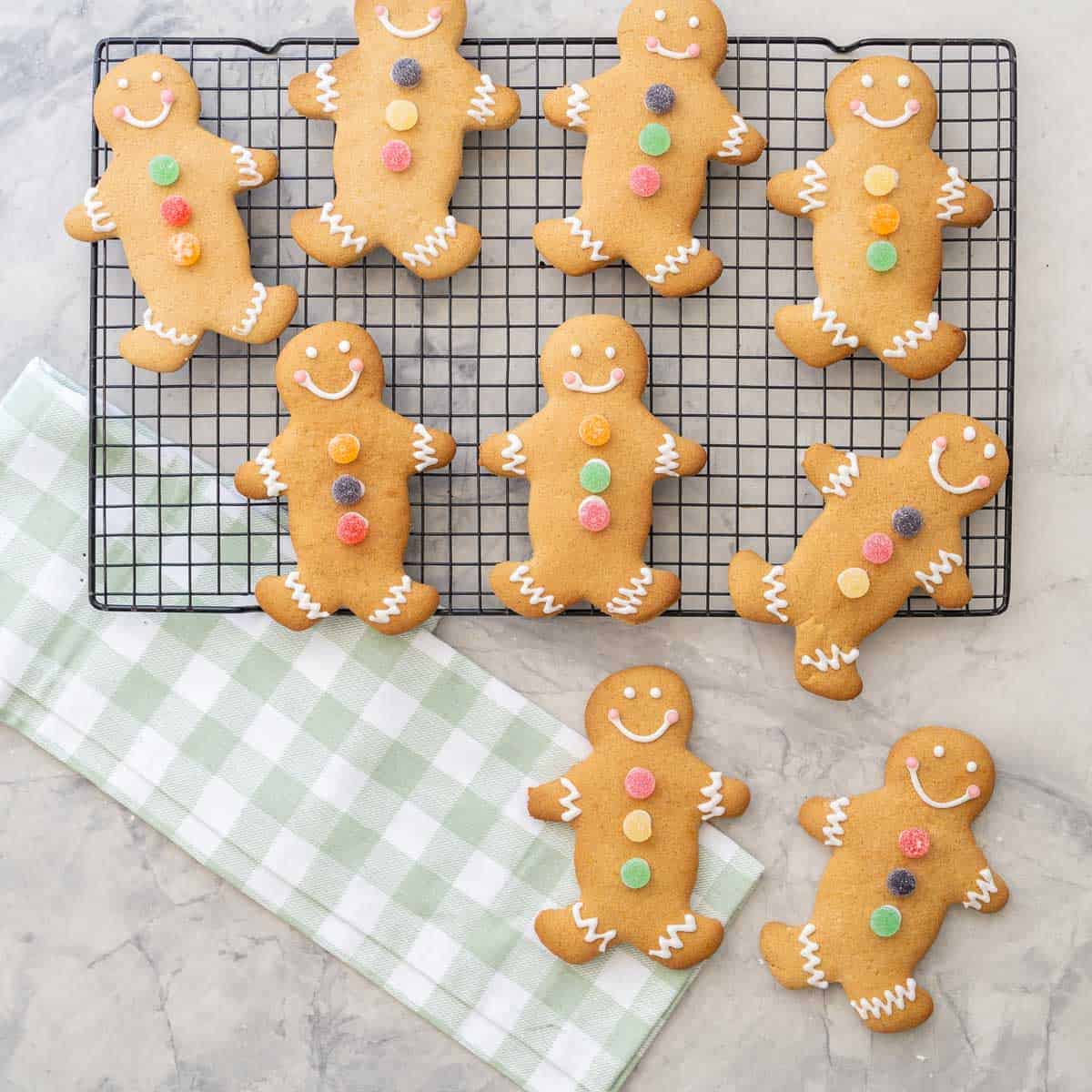 Three decorated gingerbread on cooling tray and two on bench top with green gingham checkered tea towel.