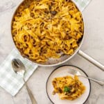 Beef bacon mushroom pasta served with a fork on dinner plate with garnish of parsley, pan of pasta on top of gingham olive green checkered tea towel with serving spoon on benchtop.