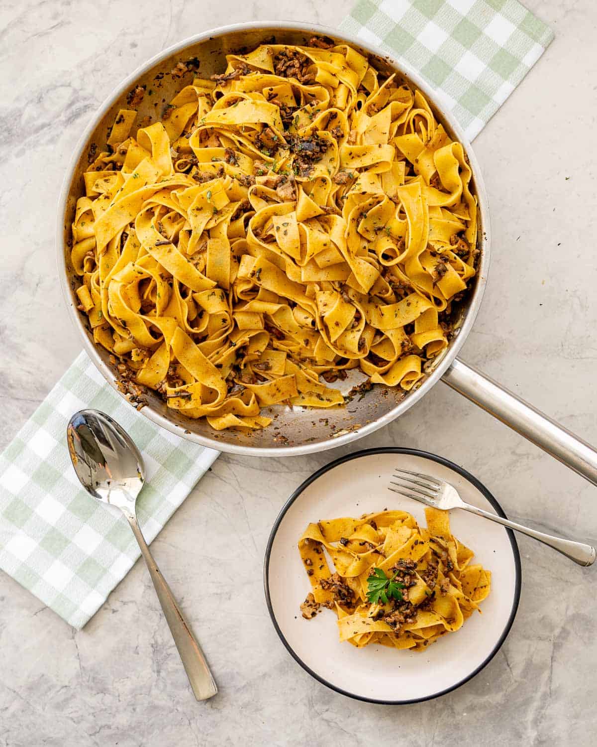 Beef bacon mushroom pasta served with a fork on dinner plate with garnish of parsley, pan of pasta on top of gingham olive green checkered tea towel with serving spoon on benchtop.