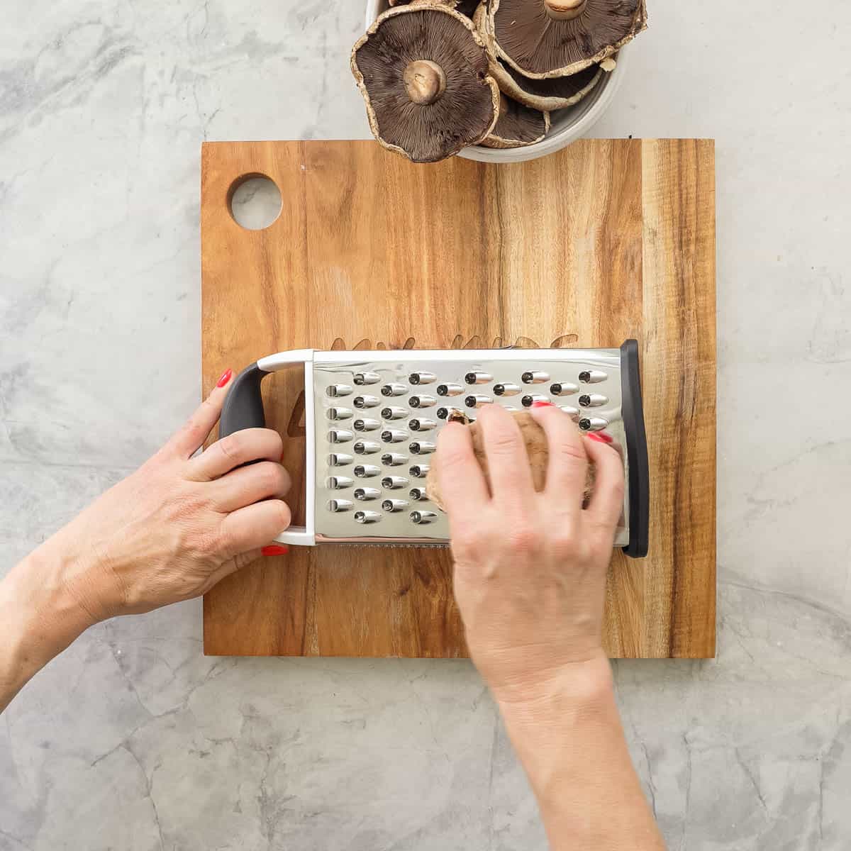 Hand holding a grater laid on its side and other hand grating a mushroom, bowl of mushrooms in bowl on benchtop.