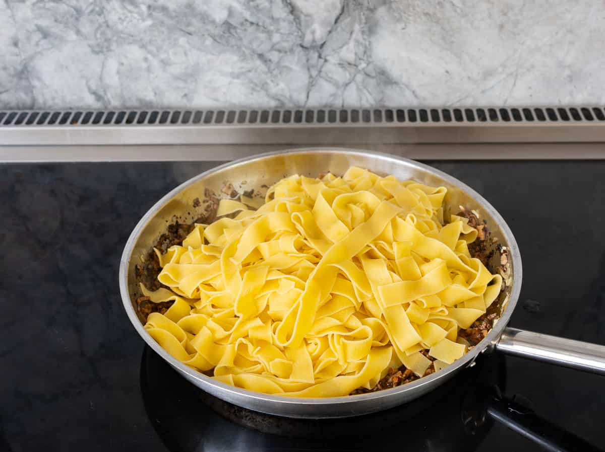 Cooked pasta added into beef mixture in frying pan on stove top.