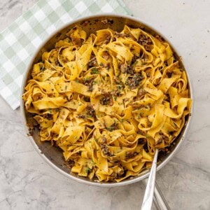 Pan of beef and bacon mushroom pasta on top of gingham olive green checkered tea towel with serving spoon inside.