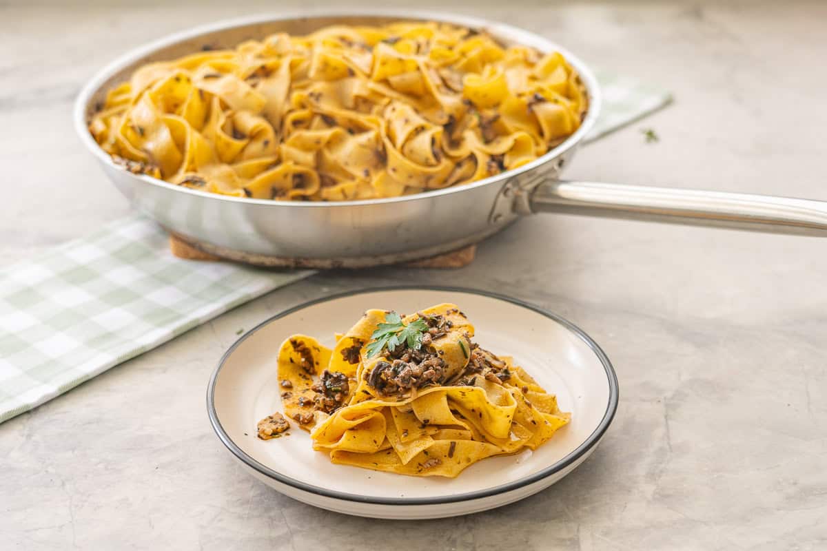 Beef bacon mushroom pasta served with a fork on dinner plate with garnish of parsley, pan of pasta on top of gingham olive green checkered tea towel with serving spoon on benchtop behind plate.