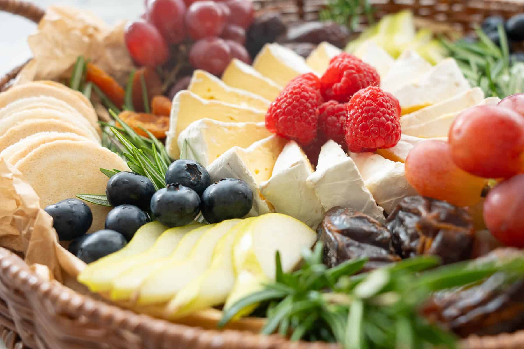 Brie cheese cut into wedges and spiralled into a circular shape, an array of different fruits, raspberries, pears, blueberry, wafer crackers, grapes, dried apricots, rosemary around the centred cheese.