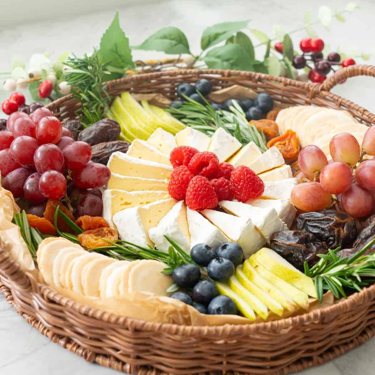 Brie cheese cut into wedges and spiralled into a circular shape, an array of different fruits, raspberries, pears, blueberry, wafer crackers, grapes, dried apricots, rosemary around the centred cheese.