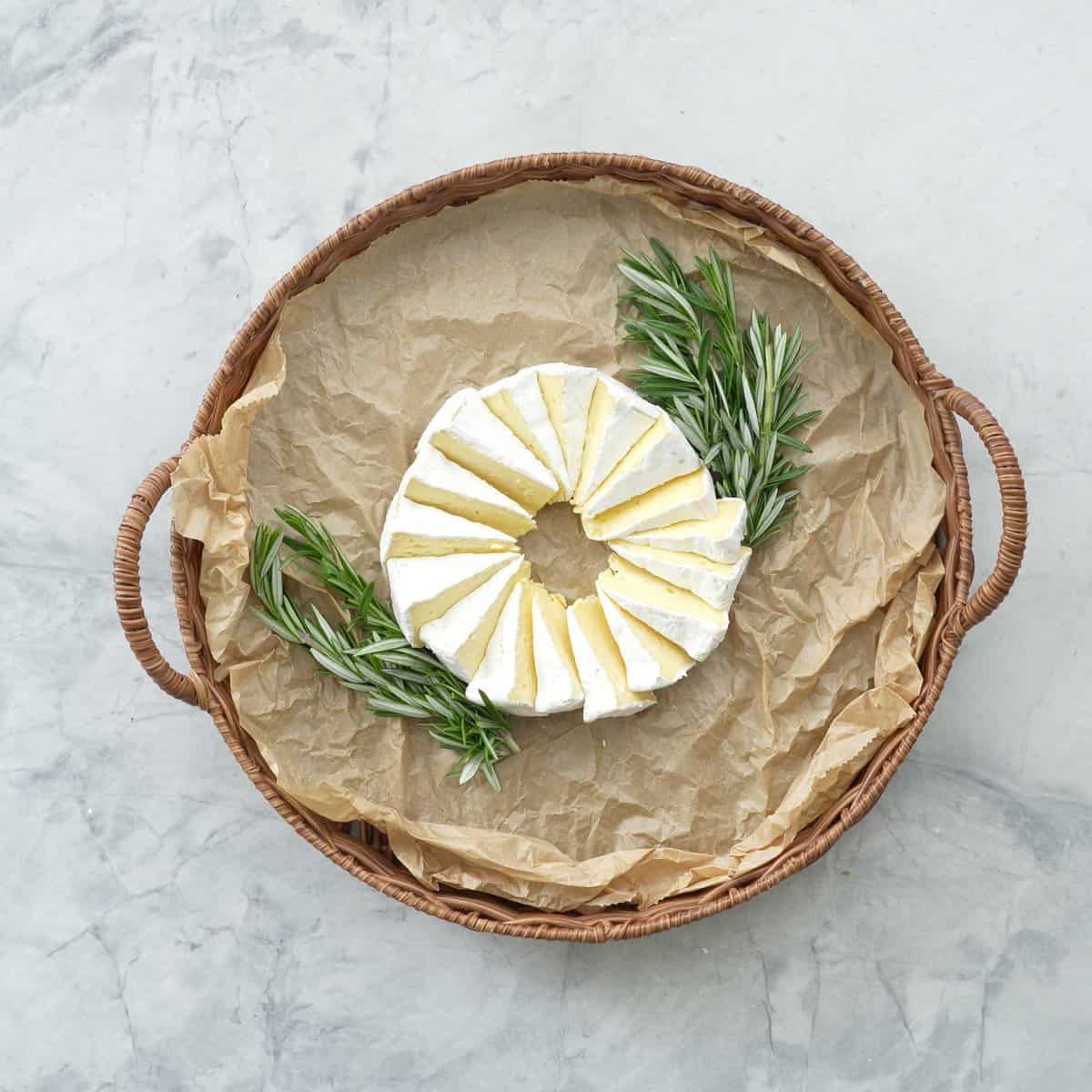 A circle platter board with Brie cheese cut into wedges and placed in the centre of board, two pieces of rosemary next to cheese.