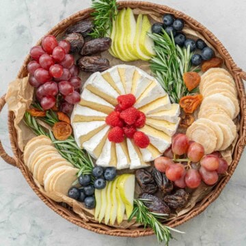 Brie cheese cut into wedges and spiralled into a circular shape, an array of different fruits, raspberries, pears, blueberry, wafer crackers, grapes, dried apricots, rosemary around the centred cheese.