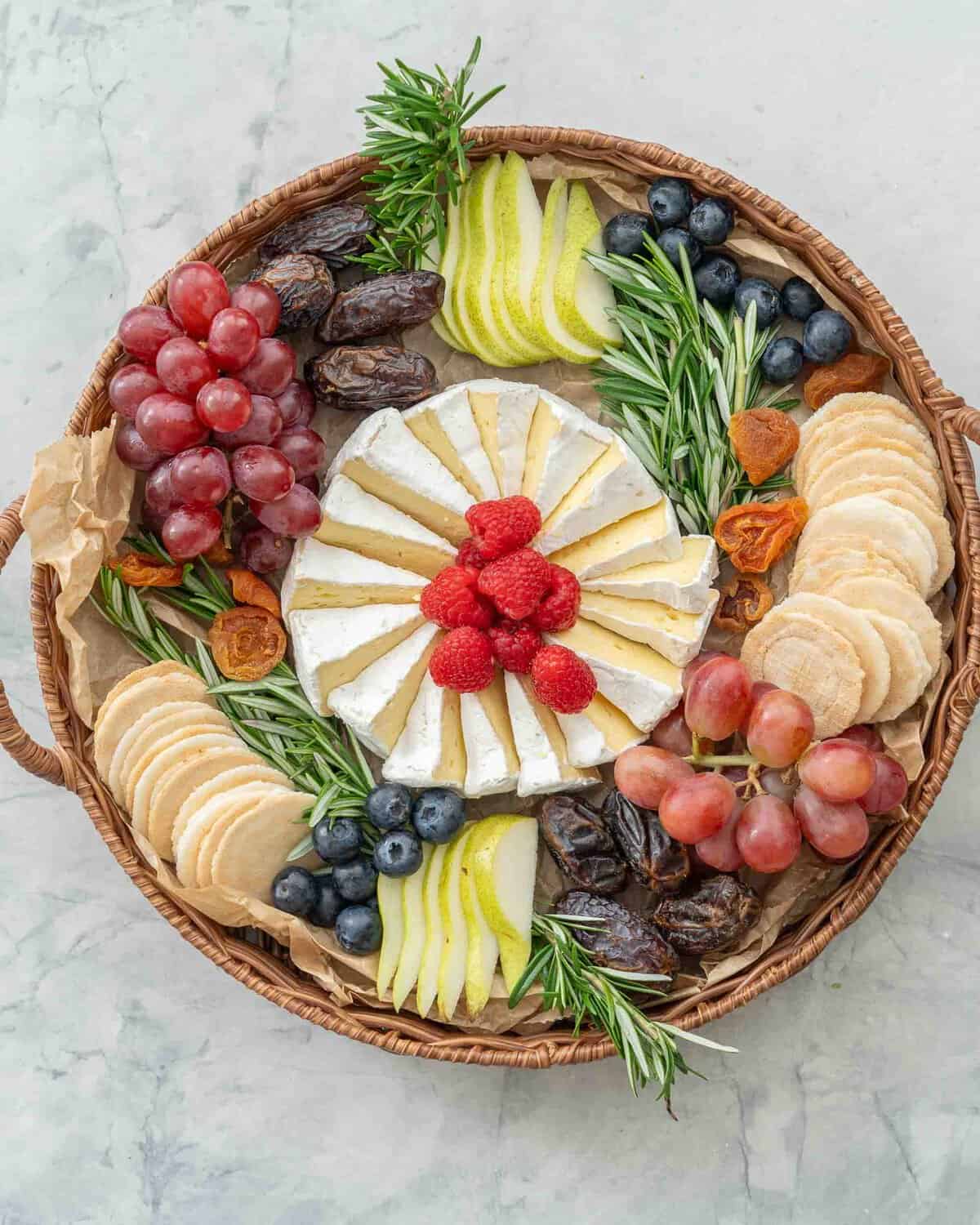 Brie cheese cut into wedges and spiralled into a circular shape, an array of different fruits, raspberries, pears, blueberry, wafer crackers, grapes, dried apricots, rosemary around the centred cheese.