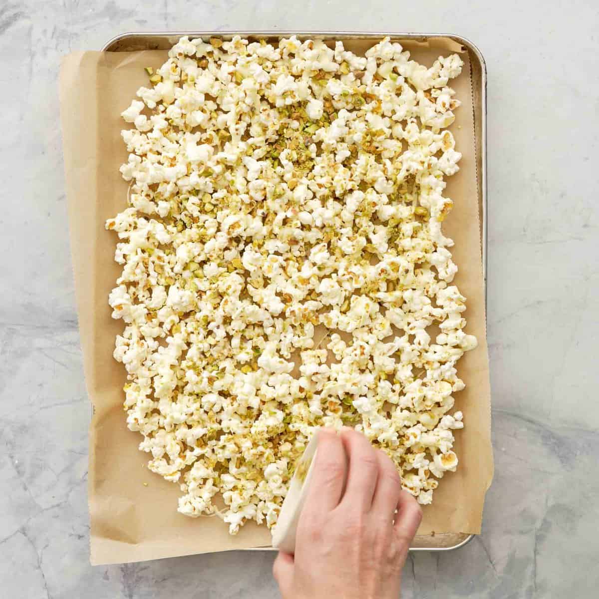 Hand sprinkling a small ceramic bowl of pistachios on top of tray of popcorn with melted white chocolate.